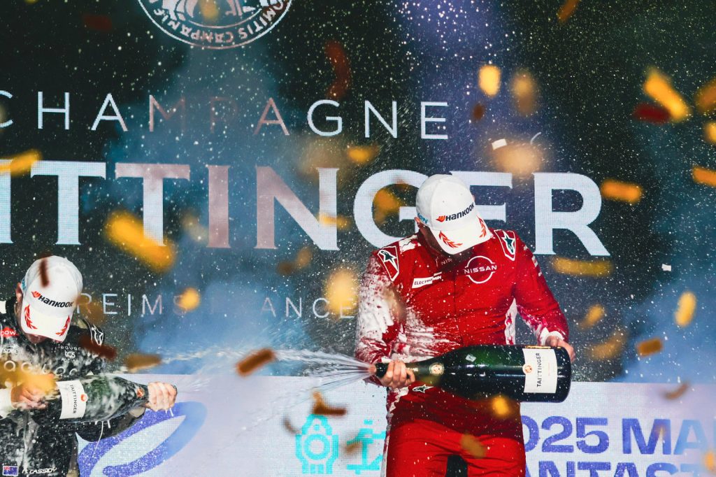 Racers in red suits spray champagne, celebrating on stage with confetti. Background features 'Taittinger' and festive atmosphere.