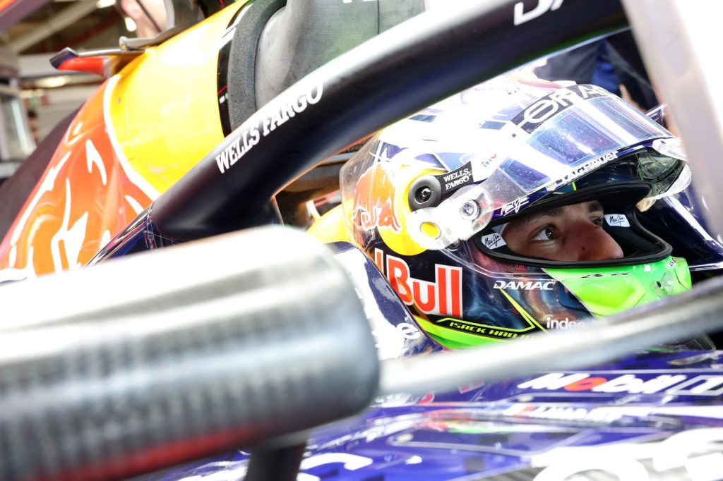 Race car driver in a colorful helmet with sponsorship logos sits focused in a Red Bull vehicle. Bright setting, intense concentration.
