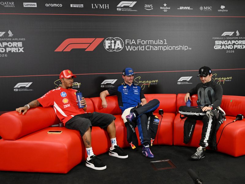 Lewis Hamilton, Max Verstappen and Charles Leclerc sit on a red sofa during a Formula 1 press conference at the 2026 Singapore Grand Prix. Hamilton wears Ferrari red, Verstappen wears Red Bull blue and Leclerc wears black Ferrari team clothing.
