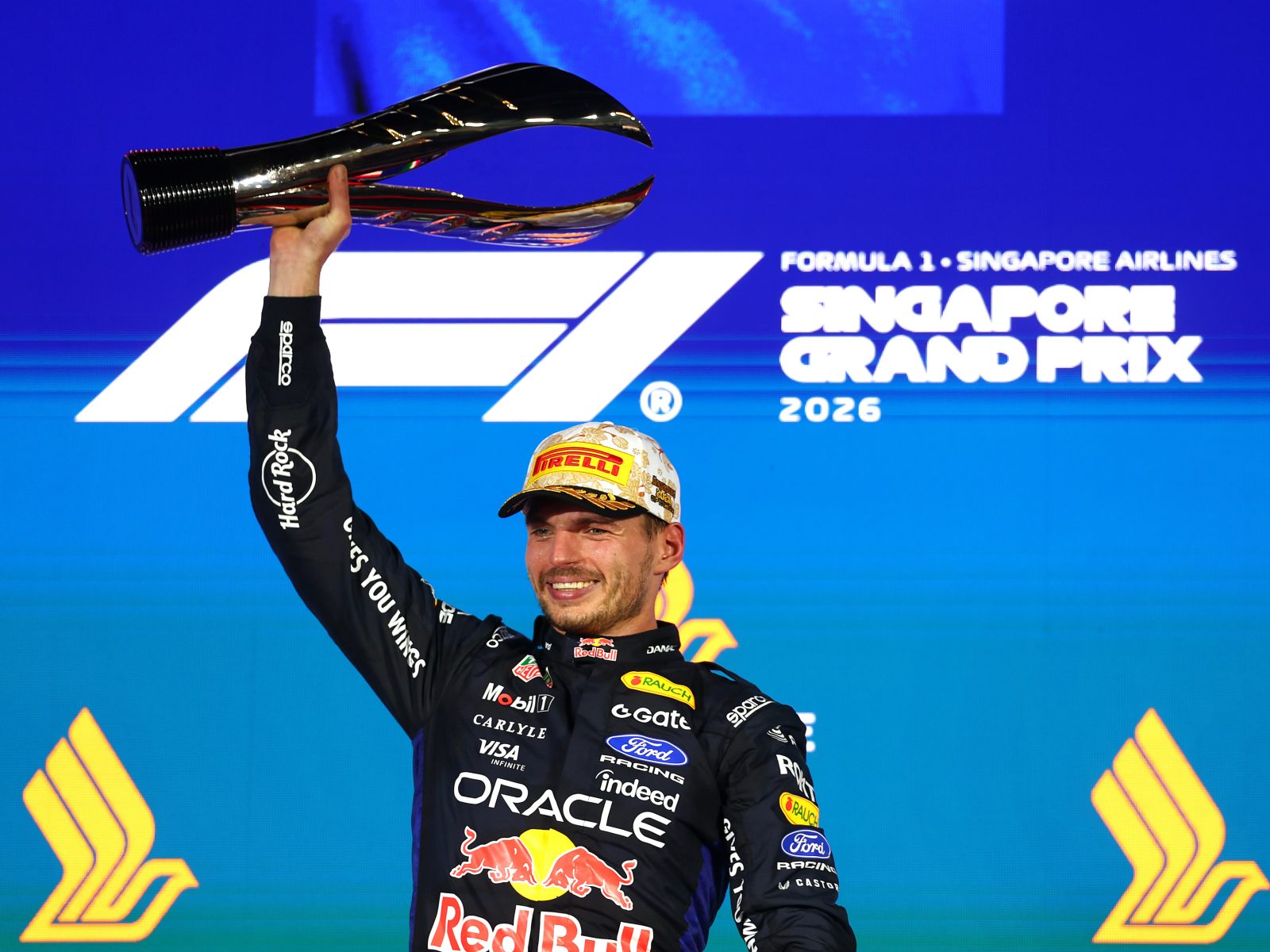 Max Verstappen smiles as he raises the Singapore Grand Prix winner's trophy above his head on the podium, wearing his Red Bull Racing suit and Pirelli cap against a blue Formula 1 Singapore Grand Prix 2026 backdrop.